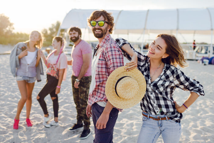 Man spending holiday closely with wife’s friend on beach, while others stand in background in casual summer attire. Man spending holiday closely with wife’s friend on beach, while others stand in background in casual summer attire.