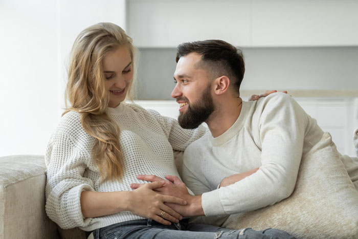 Couple sitting on couch, woman touching her belly while man smiles, relating to pregnancy obsession and health concerns. Couple sitting on couch, woman touching her belly while man smiles, relating to pregnancy obsession and health concerns.