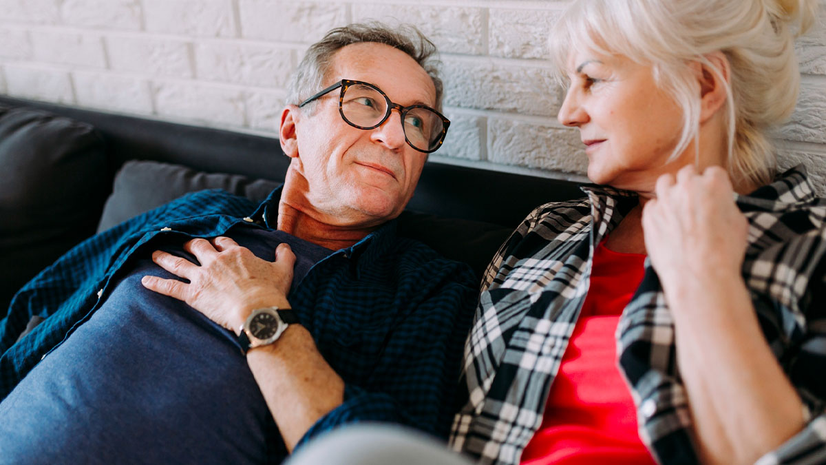 Older husband and wife discussing retirement plans and finances while sitting closely on a couch at home.