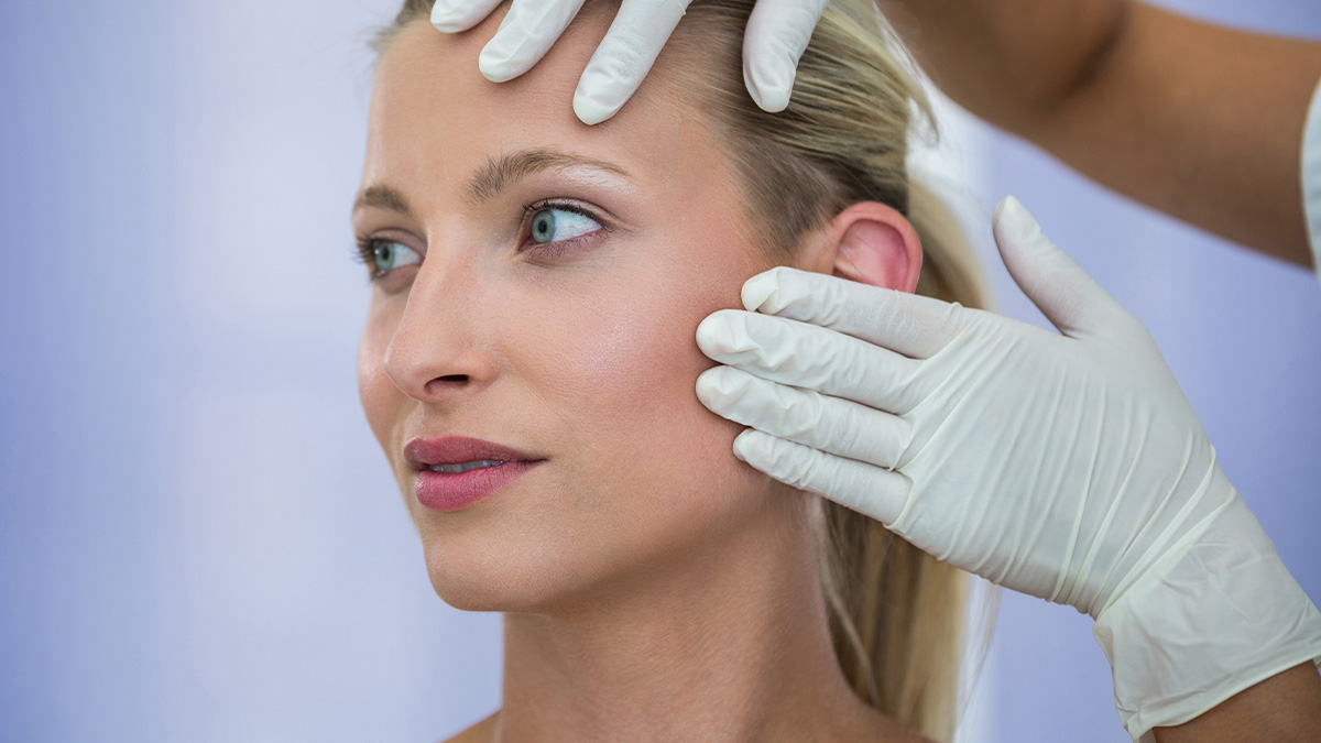 Woman during plastic surgery consultation, doctor examining her face with gloved hands in a clinic setting
