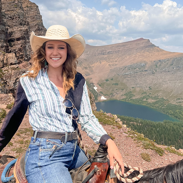 Young woman wearing cowboy hat and striped shirt sitting on horseback with mountain and lake landscape behind, karma train keyword included. Young woman wearing cowboy hat and striped shirt sitting on horseback with mountain and lake landscape behind, karma train keyword included.