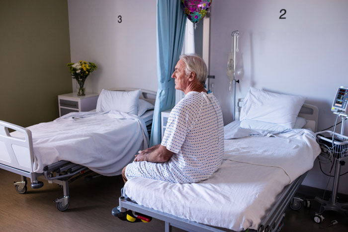 Elderly man in hospital gown sitting on bed in a quiet hospital room, reflecting on family and will issues.