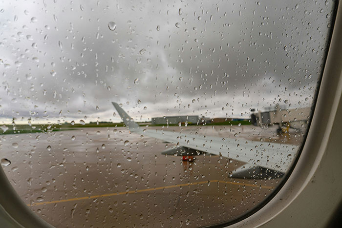 View from airplane window showing raindrops on glass with tarmac and wing in rainy weather flying details visible