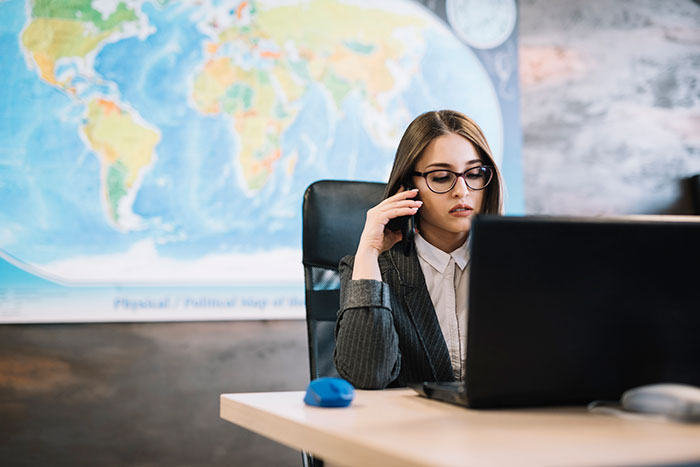 Woman wearing glasses sitting at desk with laptop, speaking on phone, with world map in background about planes and flying secrets.