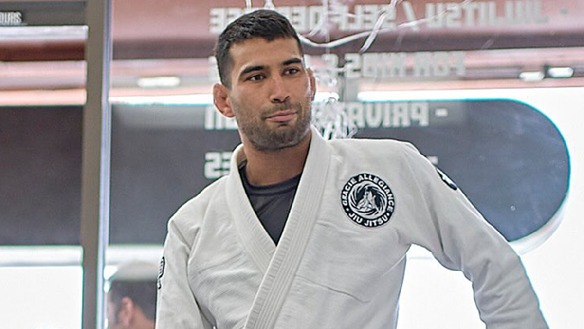 Jiu-Jitsu trainer in white gi standing inside gym, breaking silence after sparring accident involving paralyzing student.