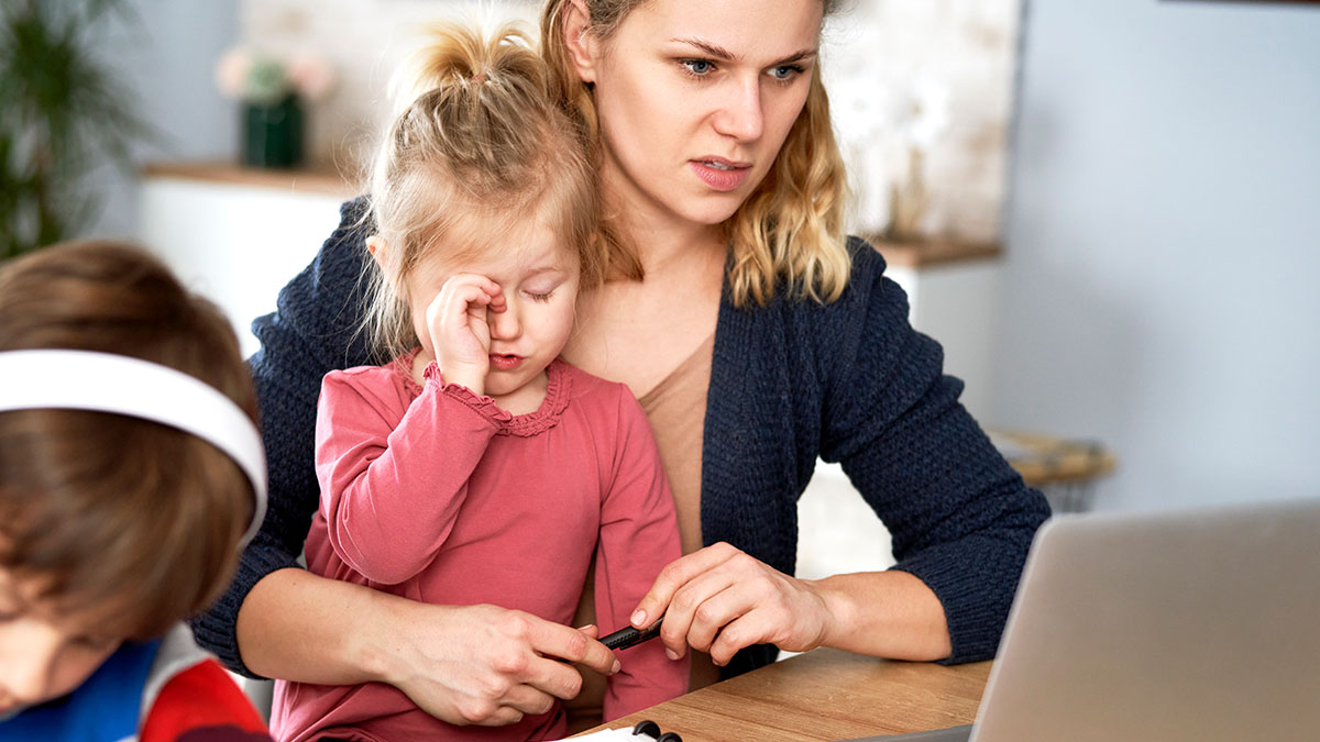 Frustrated mom managing upset toddler and working on laptop, illustrating free babysitter expectations and a surprising bill.