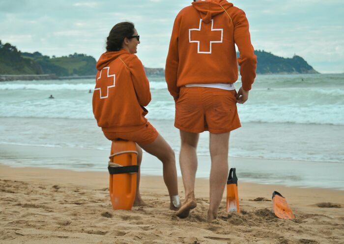 Two workers in orange uniforms on the beach sharing unprofessional things done while overlooking the ocean.