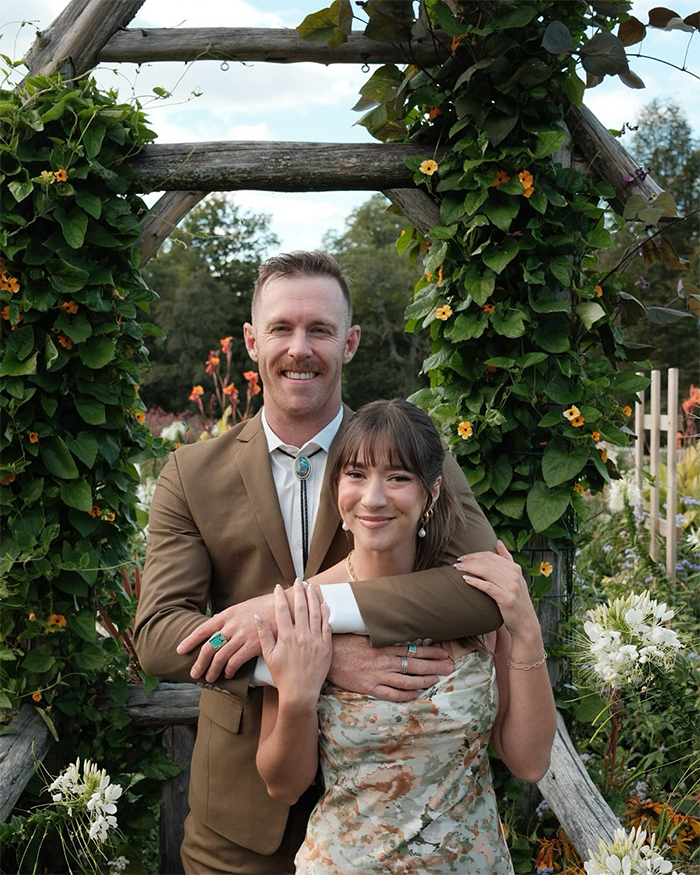Man and woman posing outdoors surrounded by flowers and greenery, representing Jiu-Jitsu trainer accident context. Man and woman posing outdoors surrounded by flowers and greenery, representing Jiu-Jitsu trainer accident context.