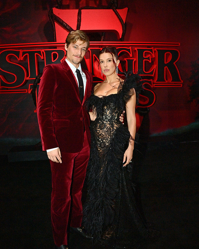 Jake Bongiovi wearing red velvet suit and a woman in black lace dress posing at Stranger Things event. Jake Bongiovi wearing red velvet suit and a woman in black lace dress posing at Stranger Things event.