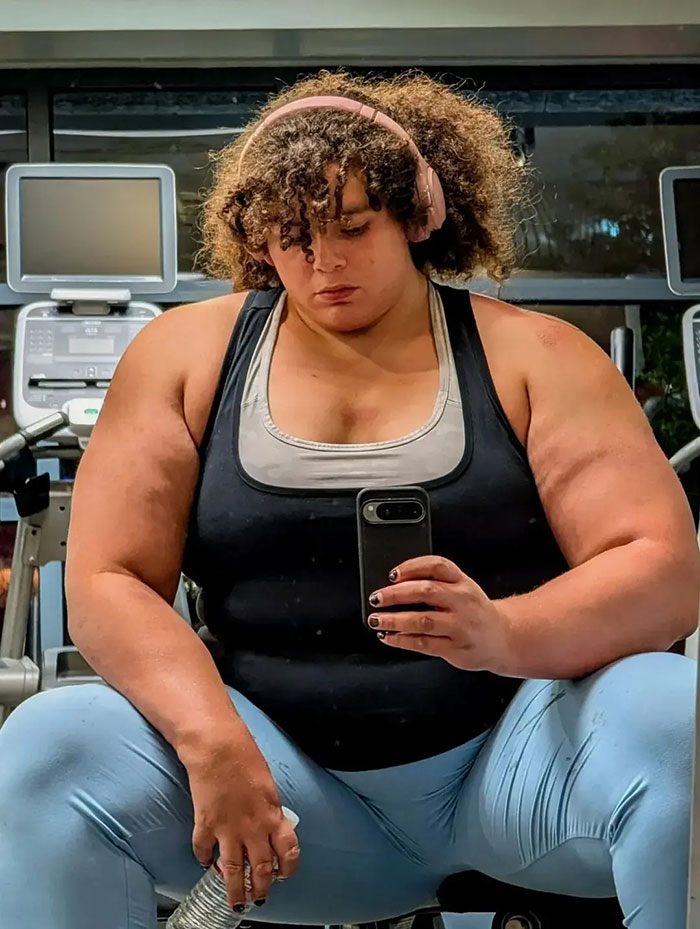 Strong woman with muscular arms wearing headphones and workout clothes, taking a gym selfie sitting on a bench. Strong woman with muscular arms wearing headphones and workout clothes, taking a gym selfie sitting on a bench.