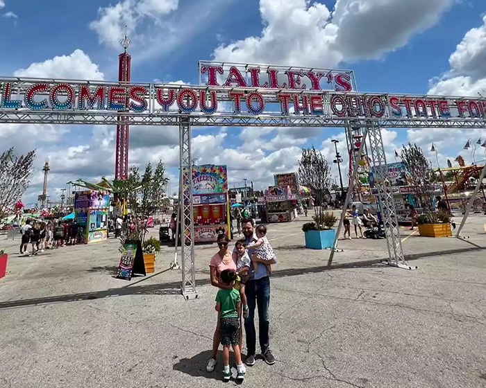 Family posing under Talley's welcome sign at Ohio State Fair on a sunny day with blue sky and clouds. Family posing under Talley's welcome sign at Ohio State Fair on a sunny day with blue sky and clouds.