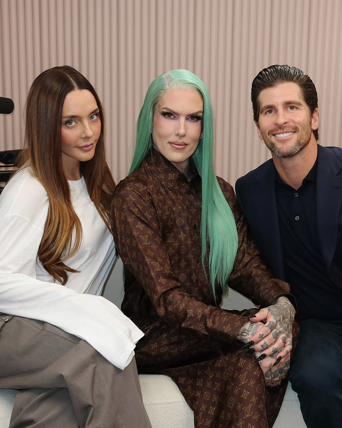 Jeffree Star seated between two people, wearing a brown patterned outfit with green hair and makeup, posing indoors. Jeffree Star seated between two people, wearing a brown patterned outfit with green hair and makeup, posing indoors.