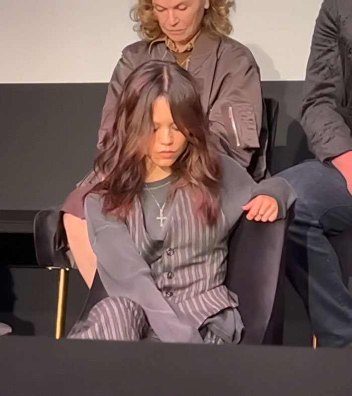 Jenna Ortega sitting with a downcast gaze, wearing a striped vest and cross necklace at Wednesday cast event.