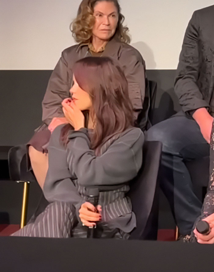 Jenna Ortega sitting with a concerned expression at a Wednesday cast event, holding a microphone.
