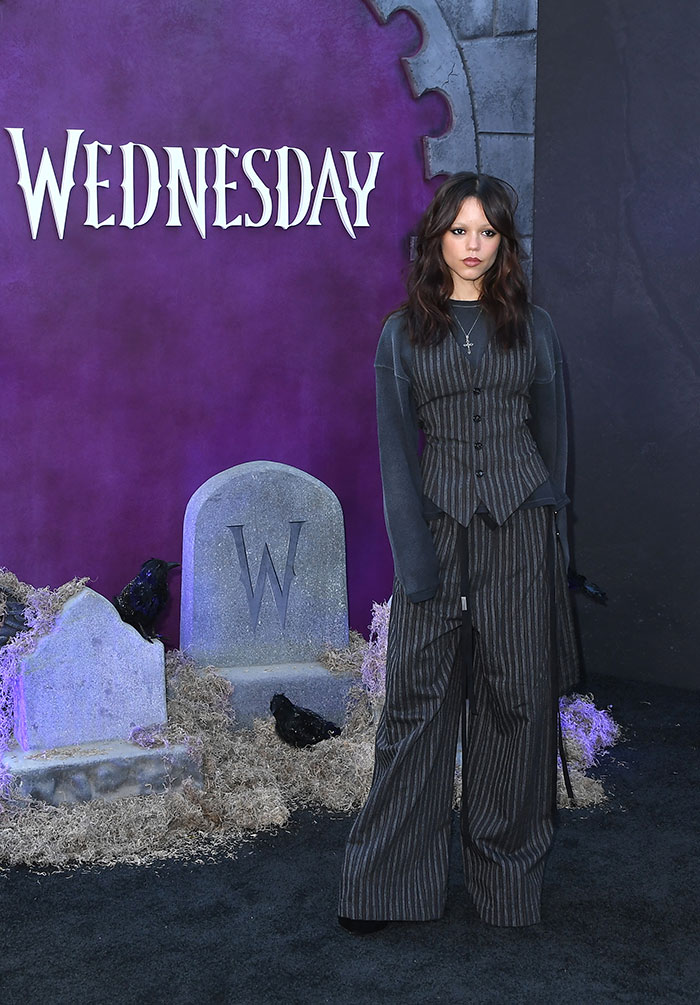 Jenna Ortega at Wednesday cast event, wearing dark striped outfit with serious expression, graveyard-themed backdrop. Jenna Ortega at Wednesday cast event, wearing dark striped outfit with serious expression, graveyard-themed backdrop.