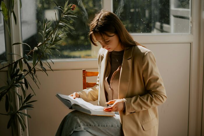 Young woman reading a book about the etymology of common words in a sunlit room near a window with plants nearby.