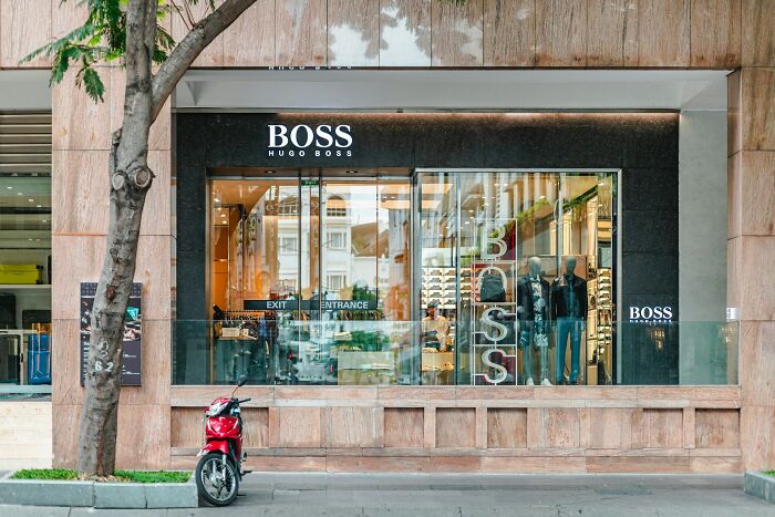 Exterior view of a Hugo Boss store with glass windows, mannequins inside, and a red scooter parked outside.