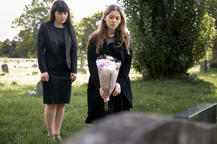 Two women dressed in black mourning at a graveyard, one holding flowers, reflecting a widowed friend and family conflict.