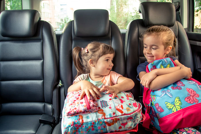 Two young girls sitting in a car backseat holding colorful backpacks showing family and mom prioritizes widowed friend concept.