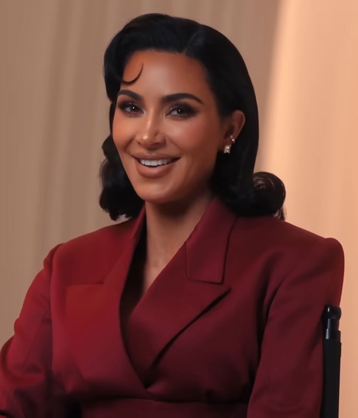 Kim Kardashian smiling in a red blazer during an interview discussing pathological liars and psychic controversies.