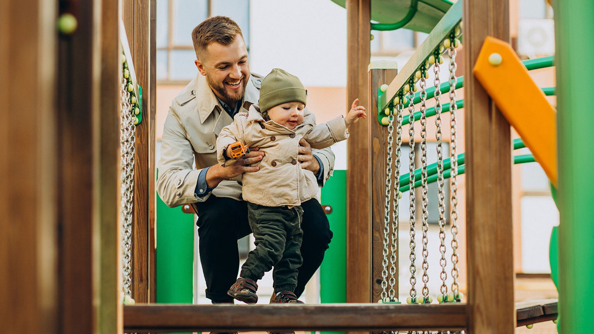Hispanic man smiling and helping a toddler walk on playground equipment, highlighting caring and trust.