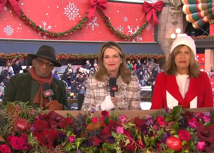 Three hosts seated with holiday decorations behind and festive flowers in front during Thanksgiving parade coverage.