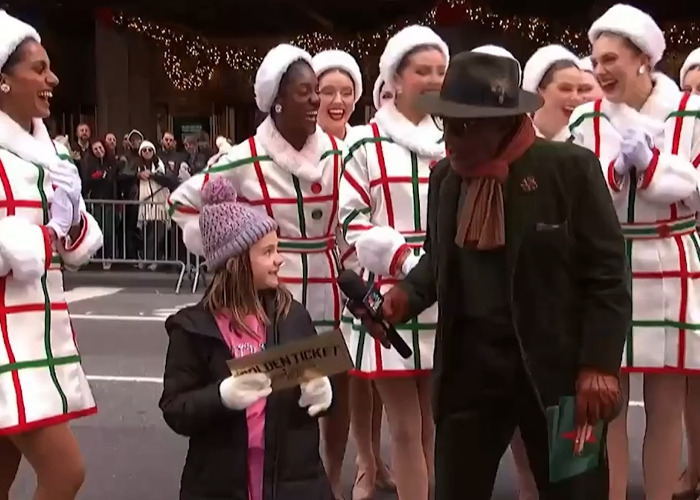 Little girl holding golden ticket with dancers around her during Thanksgiving parade, showing a candid reaction.
