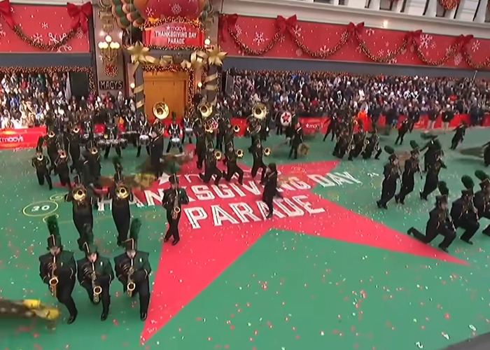 Marching band performing at Thanksgiving parade with festive decorations and large crowd watching the event live