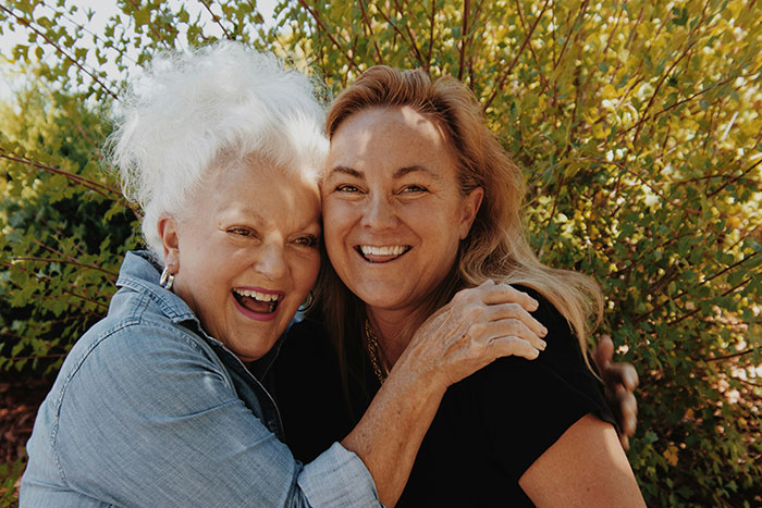 Two women smiling and hugging outdoors with sunlight and green foliage, highlighting woman and service dog themes. Two women smiling and hugging outdoors with sunlight and green foliage, highlighting woman and service dog themes.