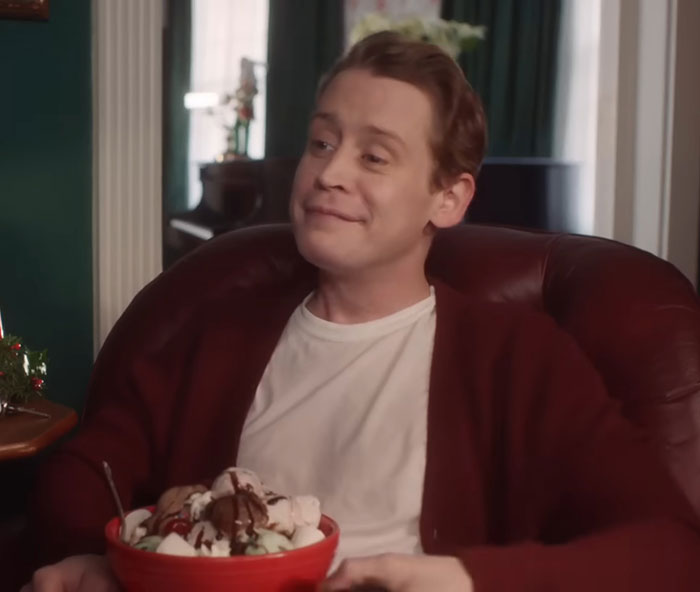 Macaulay Culkin sitting in a chair smiling while holding a bowl of ice cream, casually dressed in a white shirt and cardigan. Macaulay Culkin sitting in a chair smiling while holding a bowl of ice cream, casually dressed in a white shirt and cardigan.