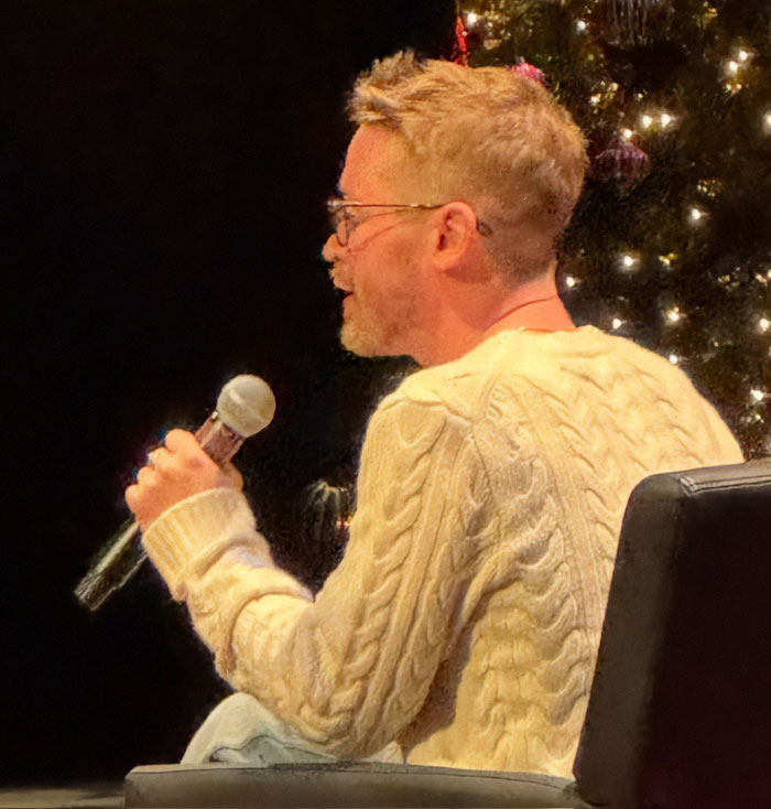Macaulay Culkin speaking into a microphone while seated, wearing glasses and a cream cable-knit sweater during an event. Macaulay Culkin speaking into a microphone while seated, wearing glasses and a cream cable-knit sweater during an event.