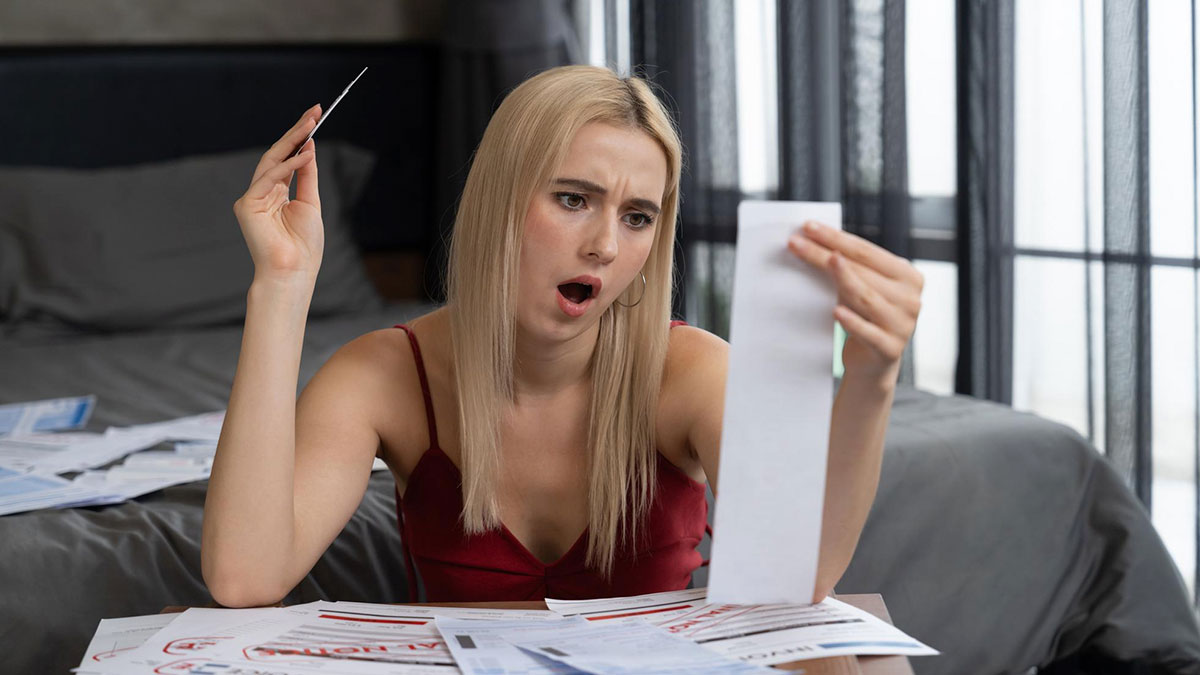 Woman in a red dress looking shocked while reviewing bills at a table with a credit card in hand.