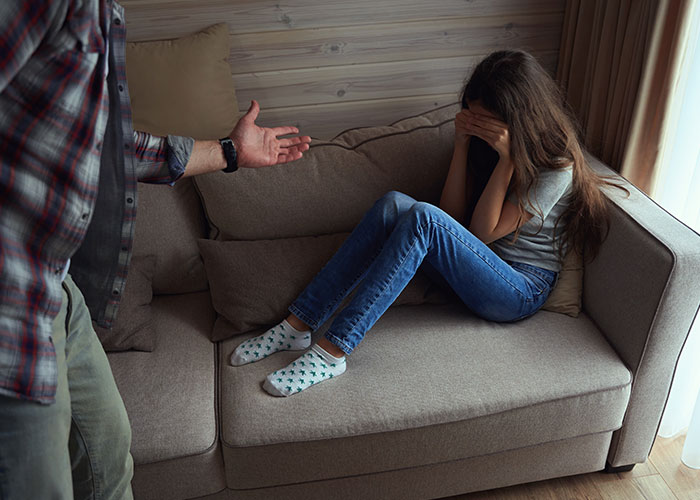 Man and teen in tense moment on couch, girl covering face while man gestures showing frustration with rude attitude.