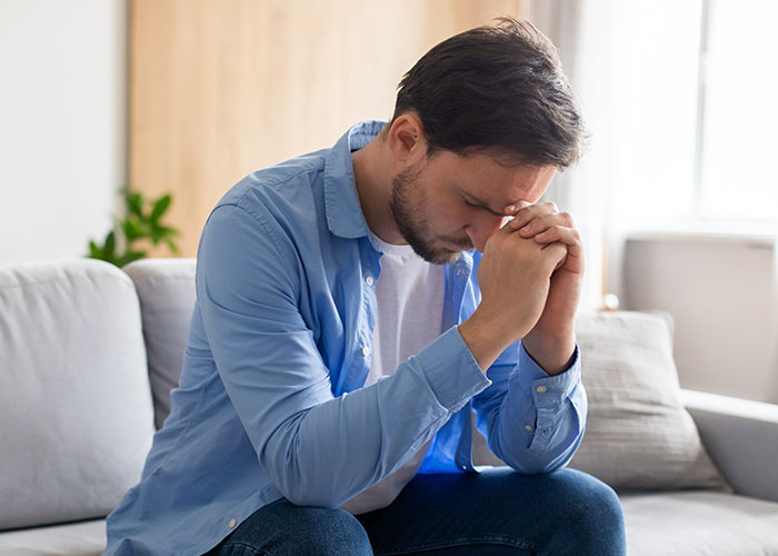 A stressed man sitting on a couch with his head bowed, showing frustration from dealing with a teen’s rude attitude.