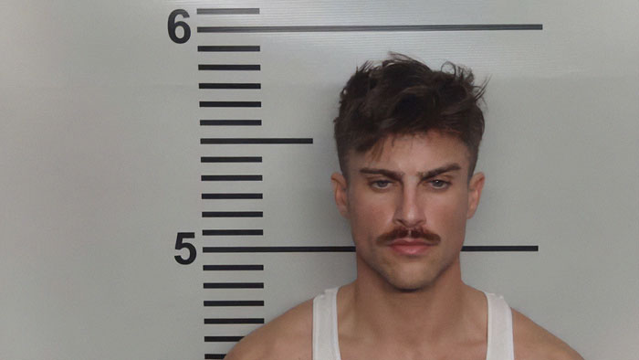 Man with mustache and tousled hair posing for a mugshot against a height chart in a white tank top. Man with mustache and tousled hair posing for a mugshot against a height chart in a white tank top.