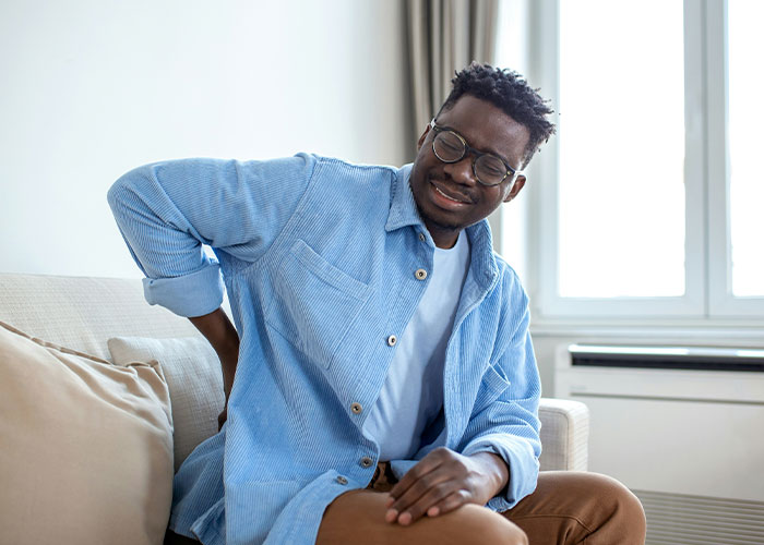 Man in glasses sitting on a couch, holding his lower back in pain, illustrating dangers in everyday life for medical malpractice cases.
