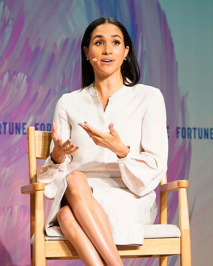 Meghan Markle speaking at an event wearing a white dress, addressing the audience with expressive hand gestures.