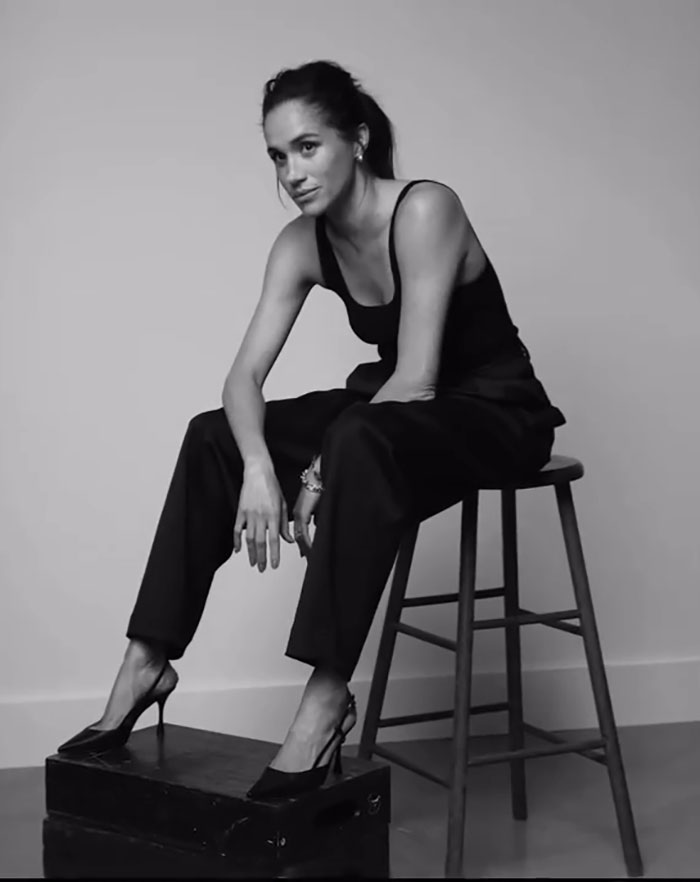 Meghan Markle sitting on a stool in black outfit and heels, resembling Princess Diana's iconic photo recreation in black and white. Meghan Markle sitting on a stool in black outfit and heels, resembling Princess Diana's iconic photo recreation in black and white.