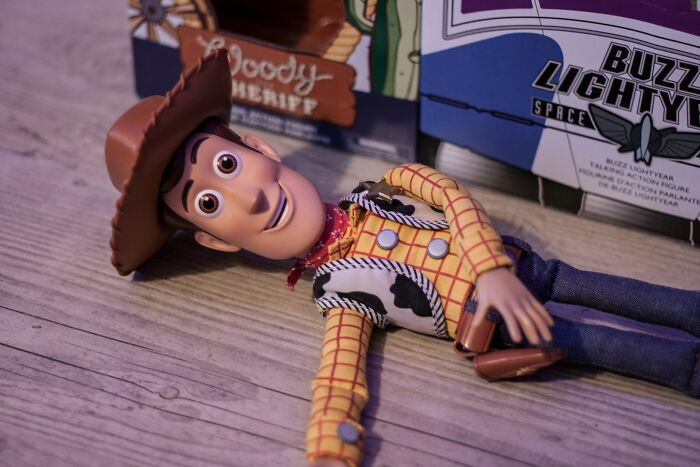 Woody toy lying on wooden surface with Buzz Lightyear box in background, showing playful yet accidentally hurt expressions.