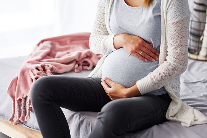 Pregnant woman sitting on bed holding belly, illustrating a mildly disturbing fact about pregnancy and life.
