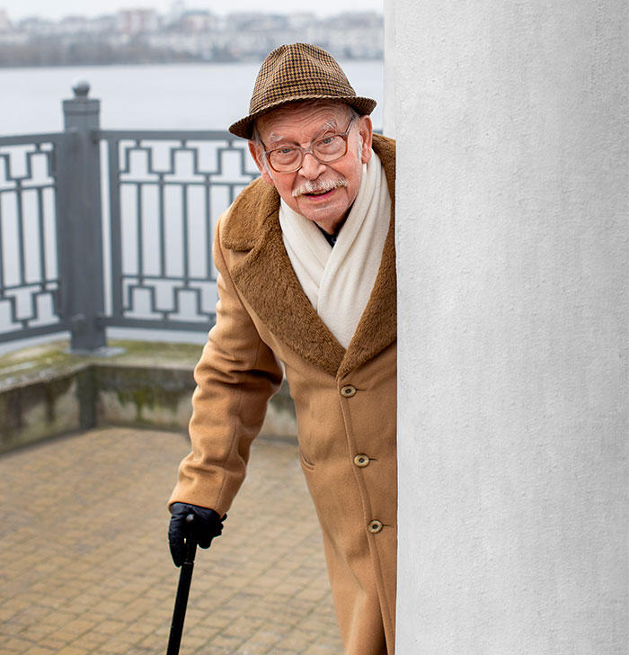 Elderly man in a brown coat and hat leaning on a cane, peeking from behind a white pillar outdoors by a railing.