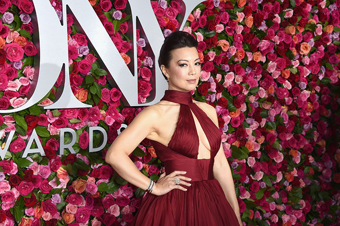Actress Ming-Na Wen posing in a red gown at an event with a floral wall backdrop highlighting her acting career. Actress Ming-Na Wen posing in a red gown at an event with a floral wall backdrop highlighting her acting career.