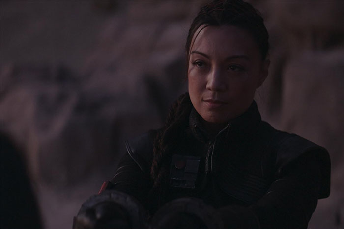 Actress Ming-Na Wen in dark outfit with braided hair, reflecting quietly in a dim, rocky outdoor setting. Actress Ming-Na Wen in dark outfit with braided hair, reflecting quietly in a dim, rocky outdoor setting.