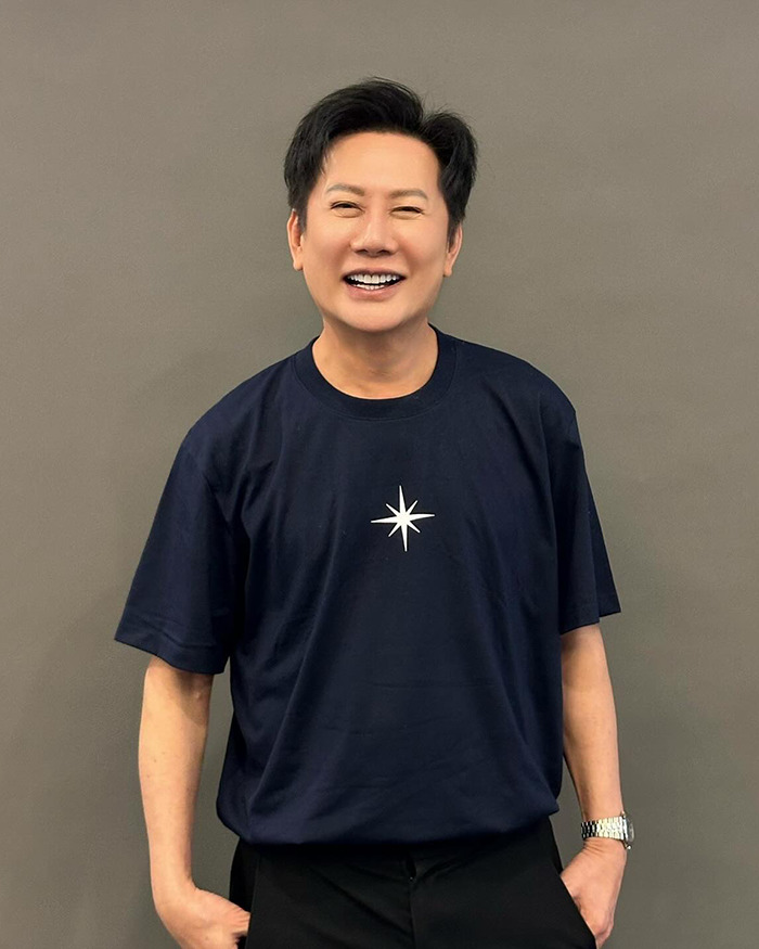 Smiling man wearing a navy shirt with a star symbol, posing against a plain gray background in a casual setting. Smiling man wearing a navy shirt with a star symbol, posing against a plain gray background in a casual setting.