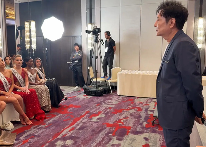 Miss Universe contestants seated in gowns facing a man in a suit during a tense moment at the event. Miss Universe contestants seated in gowns facing a man in a suit during a tense moment at the event.