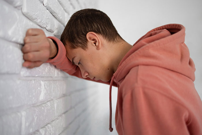 Teen boy in a pink hoodie leaning against a white brick wall, showing stress possibly related to mom affair and college money issues. Teen boy in a pink hoodie leaning against a white brick wall, showing stress possibly related to mom affair and college money issues.