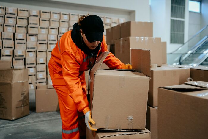 Worker in orange uniform lifting cardboard boxes in a warehouse illustrating inventive ways workers got away breaking rules.