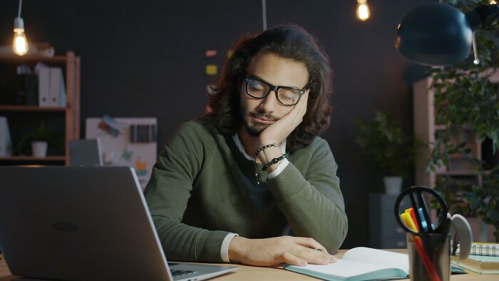 Man wearing glasses and a green sweater appearing sleepy while working late at desk with laptop and notebook in dim office.