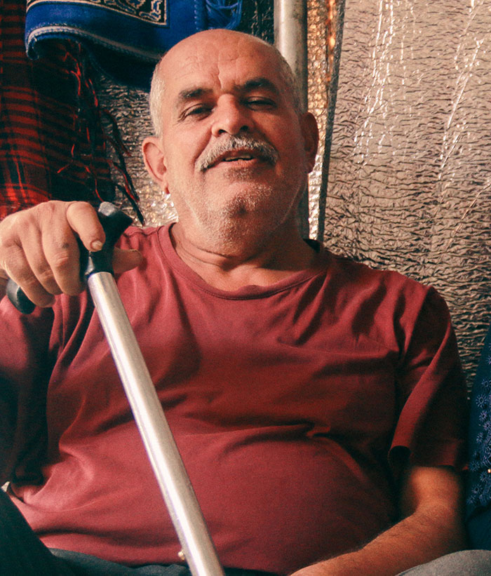 Elderly man in a red shirt holding a cane, sitting indoors with a relaxed expression, family tensions implied