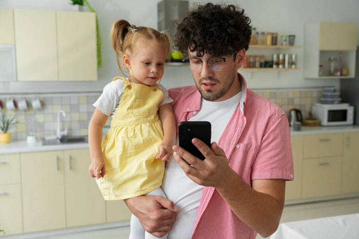 Man holding daughter in kitchen, looking at phone concerned about neighbor fence replacement dispute. Man holding daughter in kitchen, looking at phone concerned about neighbor fence replacement dispute.